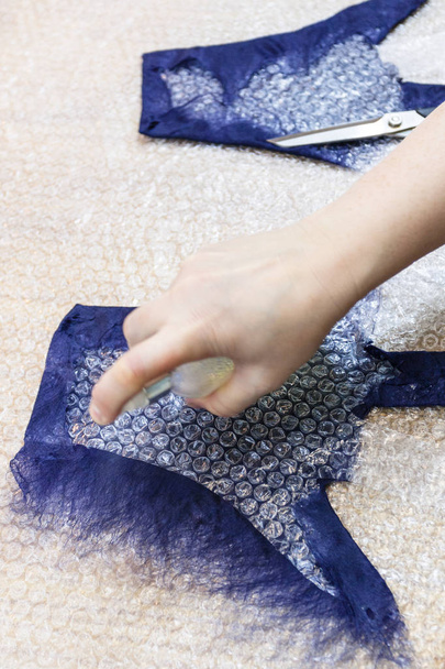 atelier van hand maken een fleece handschoenen van blauwe Merino schapenwol met behulp van NAT vilten proces - vakman bevochtiging van vezels aan achterzijde van de handschoen op snijden patroon door spray - Foto, afbeelding