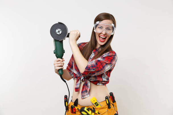 Strong young energy handyman woman in plaid top shirt, denim shorts, kit tools belt full of instruments in protective goggles hold power electric saw isolated on white background. Female in male work - Fotó, kép