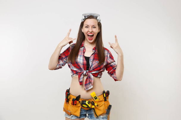 Strong young handyman woman in plaid top shirt, denim shorts, kit tools belt full of instruments in protective goggles isolated on white background. Female in male work. Copy space advertisement - Photo, Image