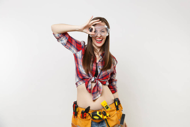Strong young handyman woman in plaid top shirt, denim shorts, kit tools belt full of instruments in protective goggles isolated on white background. Female in male work. Copy space for advertisement - Photo, Image