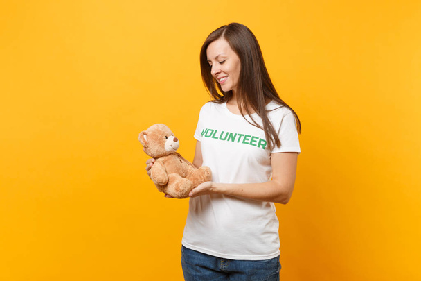 Portret van een vrouw in wit t-shirt met schriftelijke inscriptie groene titel vrijwilligers houden teddy bear knuffel geïsoleerd op gele achtergrond. Vrijwillige gratis hulp hulp, liefdadigheid gunst werk concept - Foto, afbeelding