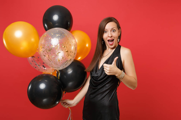Excited young woman in little black dress celebrating showing thumb up holding air balloons isolated on bright red background. St. Valentine's Day Happy New Year birthday mockup holiday party concept - Foto, imagen