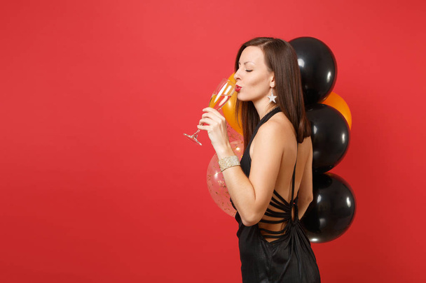 Side view of beautiful woman in black dress celebrating drinking champagne hold air balloons isolated on red background. International Women's Day Happy New Year birthday mockup holiday party concept - Фото, изображение
