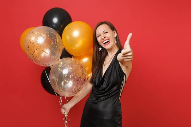 Γελώντας νεαρή γυναίκα στο μικρό μαύρο φόρεμα που δείχνει τον αντίχειρα επάνω κρατώντας αερόστατα γιορτάζει απομονώνονται σε κόκκινο φόντο. Της γυναίκας ευτυχισμένο το νέο έτος ημέρα γενεθλίων mockup holiday κόμμα έννοια - Φωτογραφία, εικόνα