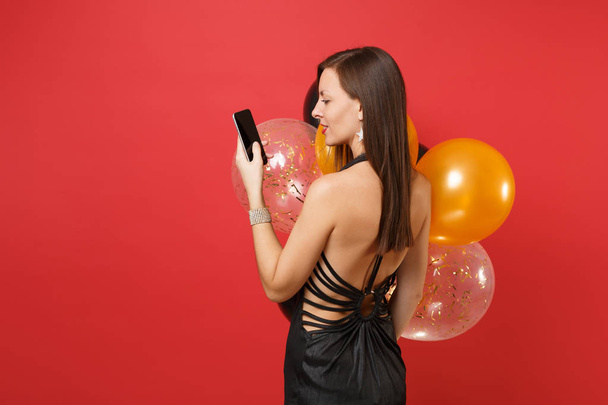 Back rear view of young woman in black dress holding, using mobile phone with blank black empty screen, air balloons isolated on red background. Happy New Year, birthday mockup holiday party concept - Zdjęcie, obraz