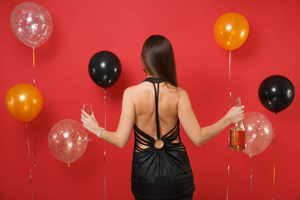 Back rear view of young girl in black dress celebrating, holding glass, bottle of champagne on bright red background air balloons. Women's Day, Happy New Year, birthday mockup holiday party concept - Photo, Image