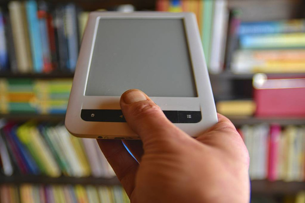 Mano sosteniendo e-book. Estantes de biblioteca en segundo plano, concepto de e-learning. Libro electrónico en mano de los hombres
 - Foto, Imagen