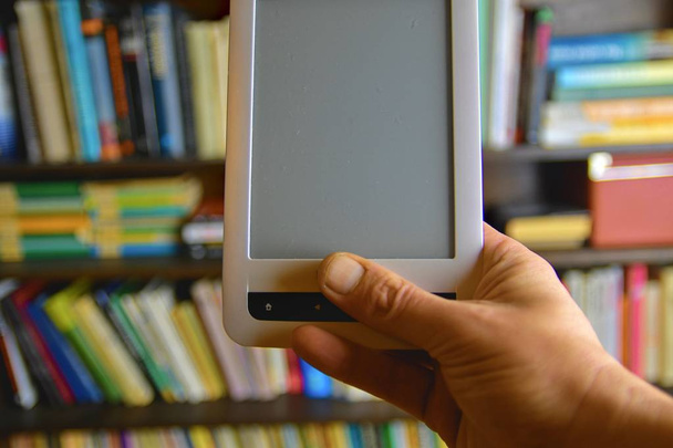 Рука держит электронную книгу. Библиотечные полки в фоновом режиме, концепция электронного обучения. Электронная книга в руках мужчин
 - Фото, изображение