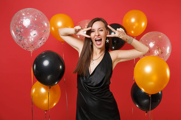 Веселая молодая женщина в маленьком черном платье празднует, показывая знак победы на ярко-красном фоне воздушных шаров. St. Valentine 's, Women' s Day, Happy New Year, birthday mockup holiday party concept
 - Фото, изображение