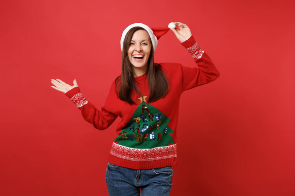 Joyful young Santa girl in knitted sweater keeping hand on Christmas hat pointing hand aside isolated on red wall background. Happy New Year 2019 celebration holiday party concept. Mock up copy space - Photo, Image