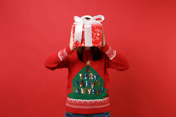 Young Santa girl in knitted sweater hiding, covering face with red box with gift present in hands isolated on red background. Happy New Year 2019 celebration holiday party concept. Mock up copy space - Фото, зображення