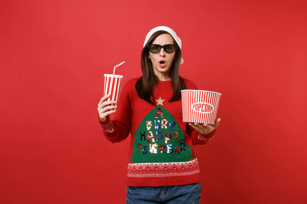 Concerned young Santa girl in 3d imax glasses watching movie film holding popcorn, cup of soda isolated on red background. Happy New Year 2019 celebration holiday party concept. Mock up copy space - Photo, Image