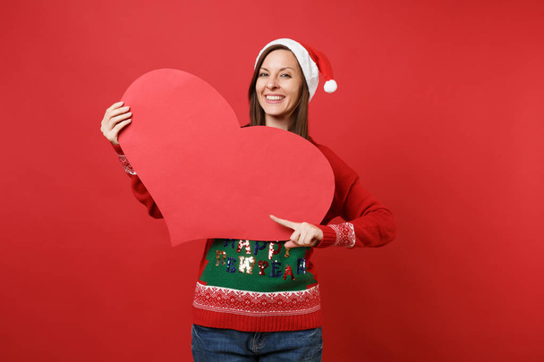 Smiling young Santa girl in Christmas hat pointing index finger on empty blank red heart in hands isolated on red background. Happy New Year 2019 celebration holiday party concept. Mock up copy space - Photo, Image