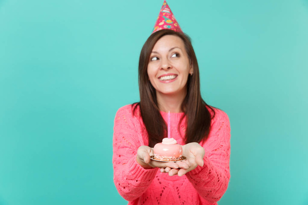 Мечтательная молодая женщина в вязаном розовом свитере и шляпе на день рождения смотрит вверх, держа в руках торт со свечой, изолированной на фоне синей стены студийного портрета. Идея образа жизни людей. Пространство для копирования
 - Фото, изображение