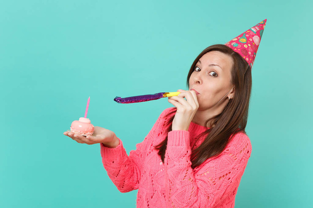 Cheerful young woman in knitted pink sweater, birthday hat with playing pipe holding in hand cake with candle isolated on blue turquoise wall background. People lifestyle concept. Mock up copy space - Photo, Image