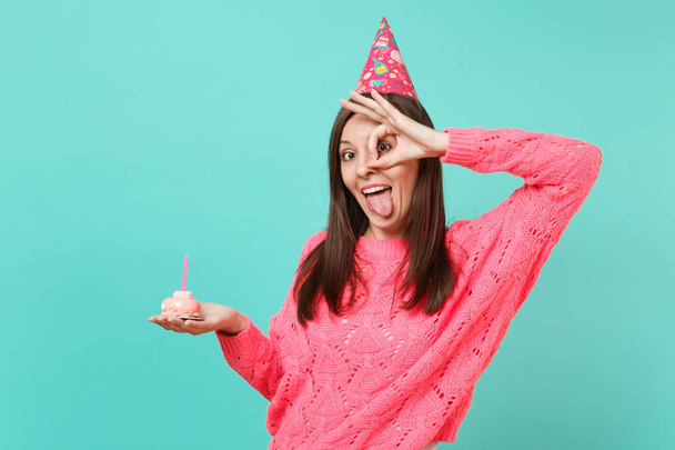 Jovem engraçada em camisola rosa de malha, chapéu de aniversário mostrando língua, gesto OK perto de olhos segurando no bolo de mão com vela isolada no fundo azul. Conceito de estilo de vida. Mock up espaço de cópia
 - Foto, Imagem