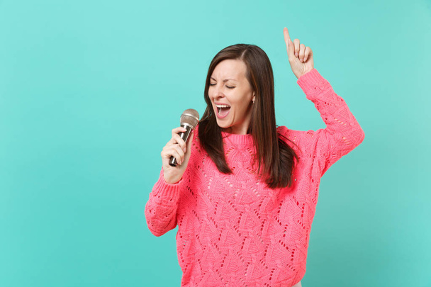 Pretty young woman in knitted pink sweater dancing, pointing index finger up, sing song in microphone isolated on blue wall background, studio portrait. People lifestyle concept. Mock up copy space - Photo, Image