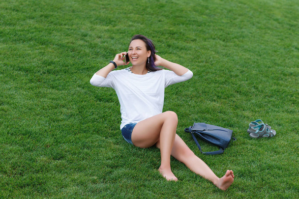Happy beautiful young woman sitting on grass and using smartphone - Fotoğraf, Görsel