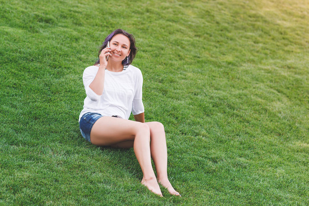 Happy beautiful young woman sitting on grass and using smartphone - Foto, imagen