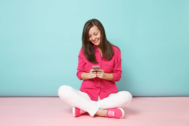 Täyspitkä muotokuva nuori hauska nainen ruusupaita valkoiset housut istua lattialla pidä matkapuhelin eristetty kirkkaan vaaleanpunainen sininen pastelli seinään tausta studio. Muoti elämäntapa käsite. Mallintaa kopiotilaa
 - Valokuva, kuva