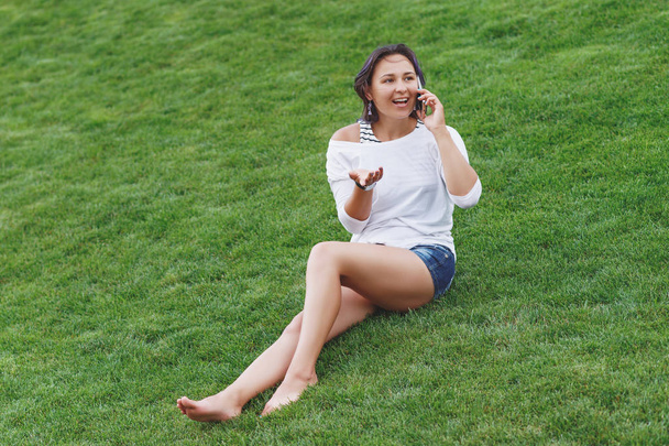 Happy beautiful young woman sitting on grass and using smartphone - Photo, Image