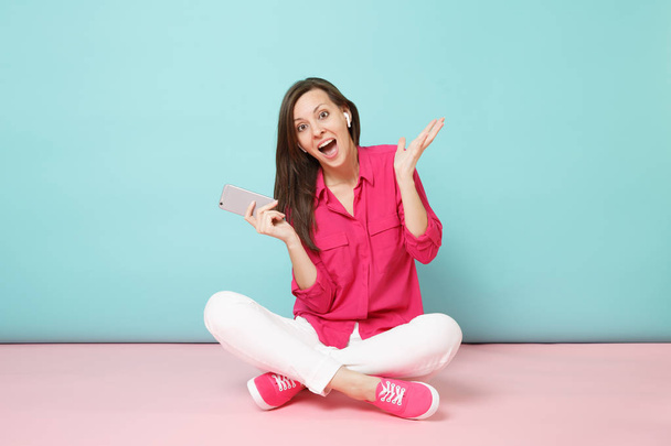 Ganzkörperporträt Frau in rosa Hemdbluse, weiße Hose auf dem Boden sitzend, Kopfhörer isoliert auf hellrosa blauer Pastellwand Hintergrundstudio. Mode-Lifestyle-Konzept. Attrappe Kopierraum - Foto, Bild