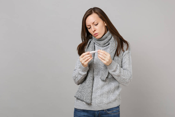 Młoda kobieta w szary sweter, szalik trzyma, patrząc na termometr na białym tle na tle szarej ścianie w studio. Zdrowy styl życia, leczenie choroby chory chory, koncepcja zimnej pory roku. Makieta do miejsca kopiowania - Zdjęcie, obraz