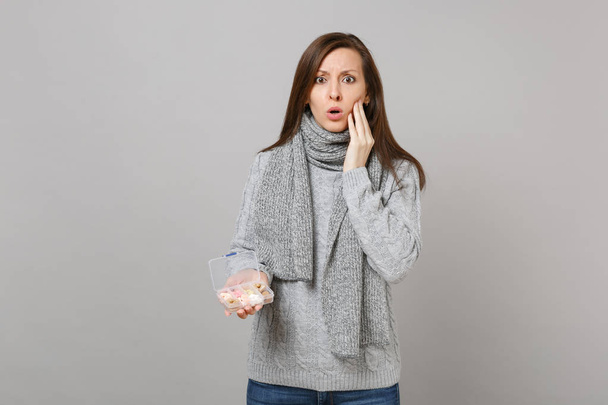 Perplexed young woman in gray sweater, scarf put hand on cheek, holding daily pill box isolated on grey wall background in studio. Healthy lifestyle, ill sick disease treatment, cold season concept - Photo, Image