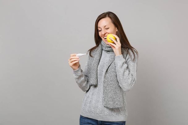 Cheerful young woman in gray sweater, scarf biting lemon, looking on thermometer isolated on grey wall background. Healthy lifestyle ill sick disease treatment cold season concept. Mock up copy space - Photo, Image