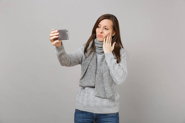 Sad young woman in sweater, scarf putting hand on cheek, doing selfie shot on mobile phone making video call isolated on grey background. Healthy fashion lifestyle people emotions cold season concept - Fotó, kép