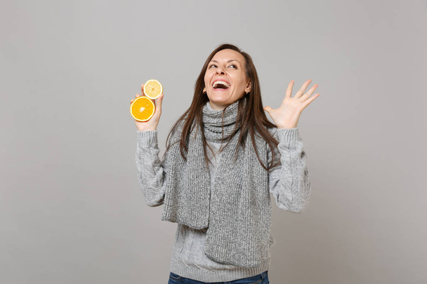 Сміється молода жінка в сірому светрі, шарф дивиться вгору, розтягує руки, тримає лимон, апельсин ізольований на сірому тлі стіни. Здоровий спосіб життя людей щирі емоції концепція холодного сезону
 - Фото, зображення