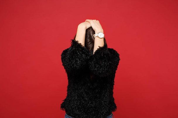 Portrait of young woman in black fur sweater putting hands on lowered head, hiding isolated on bright red wall background in studio. People sincere emotions, lifestyle concept. Mock up copy space - Photo, Image