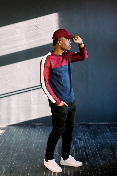 κομψό Αφρο guyportrait από μια αφρικανική-αμερικανική hipster τύπος ντυμένος με ένα κόκκινο φλις πουλόβερ και κόκκινο καπάκι στο studio. Απομονωμένα σε σκούρο φόντο με υφή φοιτητής φορώντας αθλητικά είδη - Φωτογραφία, εικόνα