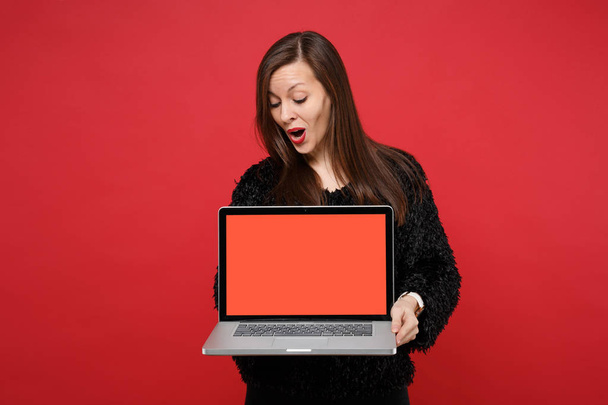 Oszołomiony młoda kobieta w swetrze czarne futro, patrząc na komputerze pc, laptopa z puste pusty ekran na białym tle na tle jasne czerwone ściany. Koncepcja życia szczere emocje ludzi. Makieta do miejsca kopiowania - Zdjęcie, obraz