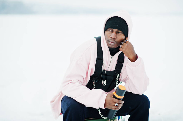 African american fisherman with thermos sitting on frozen lake. Winter fishing. - 写真・画像