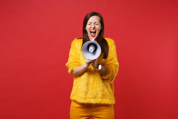 Crazy young woman in yellow fur sweater keeping eyes closed, screaming on megaphone isolated on bright red wall background in studio. People sincere emotions, lifestyle concept. Mock up copy space - Photo, Image