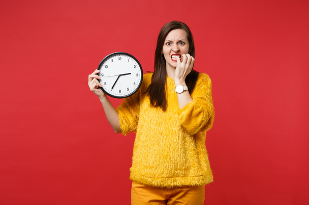 Mujer joven nerviosa en suéter de piel amarilla royendo clavos, sosteniendo reloj redondo aislado sobre fondo rojo en el estudio. El tiempo se acaba. Gente emociones sinceras, concepto de estilo de vida. Simular espacio de copia
 - Foto, imagen