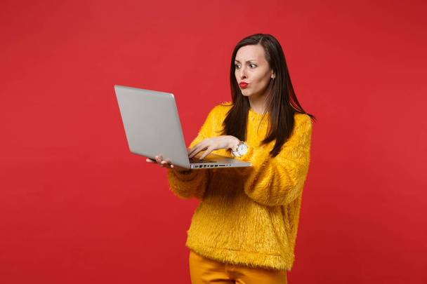 Portret van verward verbaasd jonge vrouw in geel bont trui met pc laptopcomputer geïsoleerd op heldere rode muur achtergrond in de studio. Mensen oprechte emoties levensstijl concept. Bespotten kopie databaseruimte - Foto, afbeelding