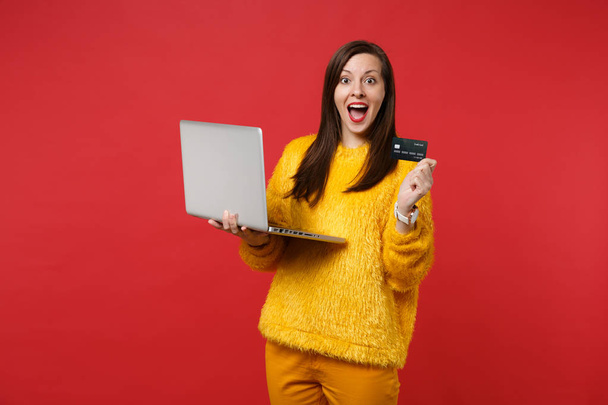 Excited young woman in yellow fur sweater keeping mouth wide open, hold credit bank card, laptop pc computer isolated on red background. People sincere emotions, lifestyle concept. Mock up copy space - Photo, Image