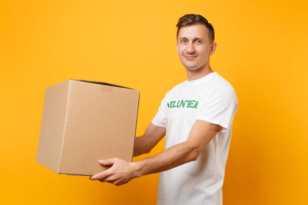Portrait of good kind man in white t-shirt with written inscription green title volunteer with big cardboard box isolated on yellow background. Voluntary free assistance help, charity grace concept - 写真・画像