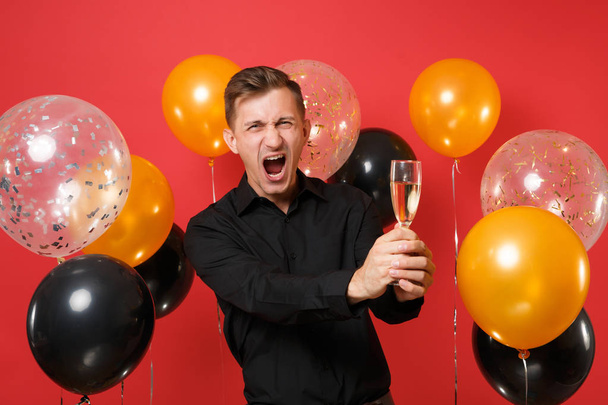 Joven loco con camisa clásica negra gritando, sosteniendo una copa de champán sobre globos de aire de fondo rojo brillante. Día Internacional de la Mujer, Feliz Año Nuevo, concepto de fiesta simulada de cumpleaños
 - Foto, Imagen