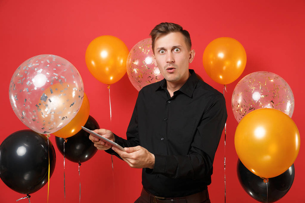 Shocked young man in black classic shirt holding and using tablet pc computer on bright red background air balloons. International Women's Day, Happy New Year, birthday mockup holiday party concept - Fotografie, Obrázek