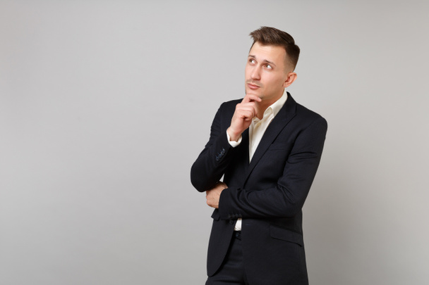 Pensive young business man in classic black suit, shirt looking up, put hand prop up on chin isolated on grey wall background in studio. Achievement career wealth business concept. Mock up copy space - Fotoğraf, Görsel