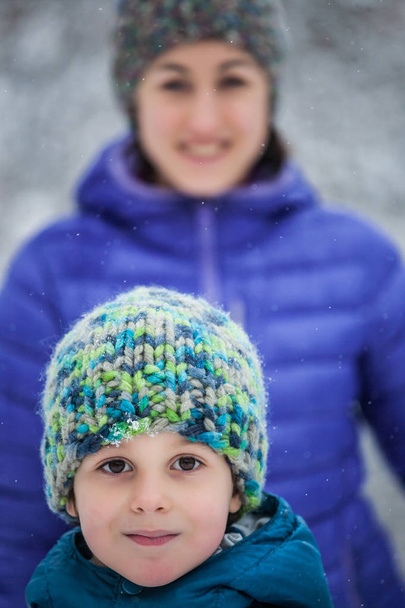 Portret van een vrouw met een kind. De jongen met zijn moeder tegen de achtergrond van de wintersneeuw bedekt bos. Gebreide mutsen. Winter wandeling. Leuke jongen. - Foto, afbeelding