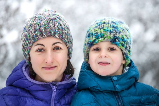 Portret van een vrouw met een kind. De jongen met zijn moeder tegen de achtergrond van de wintersneeuw bedekt bos. Gebreide mutsen. Winter wandeling. Moederdag knuffels. - Foto, afbeelding