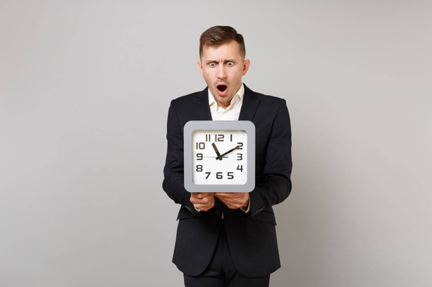 Shocked young business man in classic black suit and shirt keeping mouth wide open, looking on square clock isolated on grey background. Achievement career wealth business concept. Mock up copy space - Photo, Image