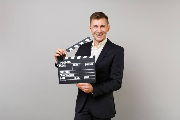 Joyful young business man in classic black suit, shirt holding classic black film making clapperboard isolated on grey wall background. Achievement career wealth business concept. Mock up copy space - Photo, Image