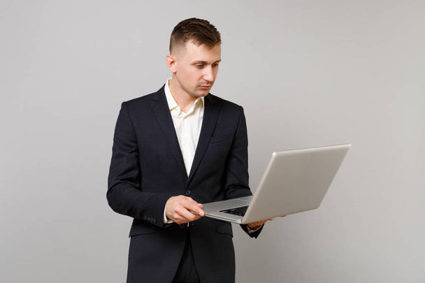 Портрет молодого бизнесмена в классическом черном костюме и рубашке, работающего на ноутбуке компьютера изолирован на сером фоне стены в студии. Достижение карьерного богатства бизнес-концепции. Пространство для копирования
 - Фото, изображение