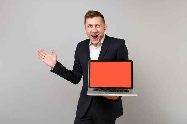Surprised young business man in suit keeping mouth wide open spreading hands holding laptop pc computer with blank empty screen isolated on grey background. Achievement career wealth business concept - Фото, изображение