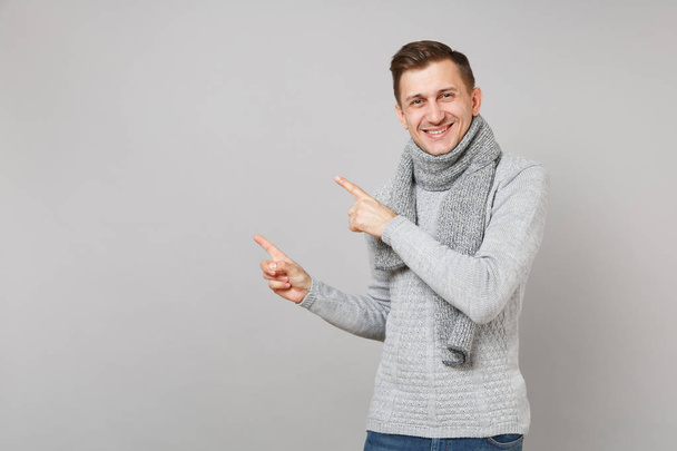 灰色のセーター、スカーフは、灰色の壁の背景に分離された脇人差し指を指しているに魅力的な若い男。健康的なファッション、ライフ スタイルの人々 の誠実な感情、寒い季節の概念。コピー スペースをモックアップします。 - 写真・画像
